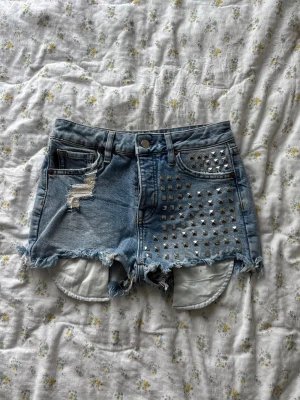 Blå jeansshorts med nitar och slitningar - Säljer ett par ljusblå jeansshorts från Superdry med råa kanter, slitningar och silverfärgade nitar på ena framsidan. Klassisk femficksmodell med hög midja och fransig nederkant. 