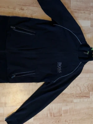 Svart kofta från Hugo Boss - Säljer en svart kofta från Hugo Boss med diskret logga på bröstet och vita detaljer längs sömmarna. Jackan har två snedställda dragkedjefickor och hög krage. Perfekt för dig som vill ha en stilren och sportig look.