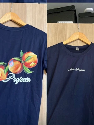 Vänster är baksida höger är fram sida. Nude project T-shirt  - Mörkblå t-shirt från nude  Projects med tryckta persikor och logga på ryggen samt liten logga framtill. Klassisk rund hals och korta ärmar. Perfekt för dig som gillar färgglada detaljer och streetwear vibes.