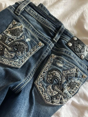 Blå miss me jeans med broderade bakfickor - Snygga blå jeans från Miss Me med detaljerade broderier och paljetter på bakfickorna. Nästan aldrig använda. Jeansens design har kontrastsömmar och slitningar som ger en cool look. Perfekt för dig som vill sticka ut med unika detaljer och stil.