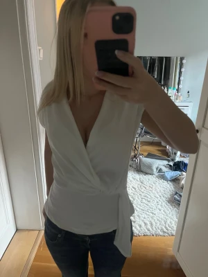 Vit omlottopp med knyt i midjan - Snygg vit omlottopp med knyt i midjan och v-ringad hals. Toppen är ärmlös och har en figurnära passform som framhäver midjan. Perfekt att styla med jeans eller kjol för en clean och fräsch look.