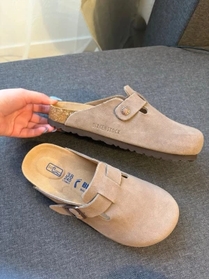 Beige Birkenstock tofflor  - Säljer ett par snygga beige tofflor från Birkenstock i mjuk mocka. De har en klassisk slip-in modell med justerbar rem och korksula för extra komfort. Perfekta för dig som gillar stilrena och bekväma skor till vardags. Priset kan diskuteras 🫶🏻