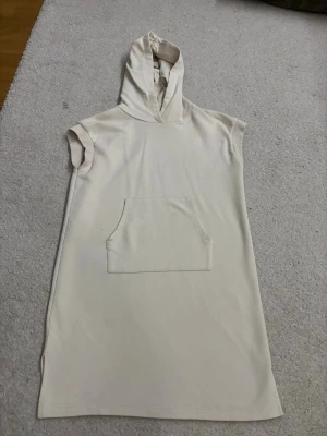 Beige ärmlös hoodieklänning - Säljer en beige, ärmlös hoodieklänning med huva och stor magficka framtill. Klänningen har en rak passform och är tillverkad i mjukt bomullstyg. Perfekt för en avslappnad och chill stil. Den har en liten fläck på magfickan men går superlätt att tvätta bort! Skicka för fler bilder!