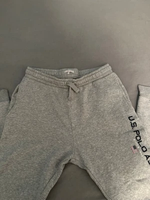 Gråa mjukisbyxor från U.S. Polo Assn. - Snygga gråa mjukisbyxor från U.S. Polo Assn. med dragsko i midjan och tryckt logga längs ena benet. Byxorna har resår i midjan och vid bensluten samt sidofickor. Perfekta för chill dagar eller träning.