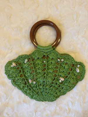 Grön virkad handväska med pärlor - Unik handväska i grön virkad design med snäckform och bruna runda handtag. Framsidan är dekorerad med pärlor och små snäckor för en cool och bohemisk look. Perfekt för dig som gillar handgjorda accessoarer och vill sticka ut.
