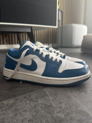 Nike Air Jordan 1 Low blå/vit - Snygga Nike Air Jordan 1 Low sneakers i blått och vitt läder. Skorna är använda och har små defekter, vilket jag tagit bild på.   Defekter:  Lite creasing vid framsidan av skon Slitage vid skosnören Lite missfärgning vid ”skotungan”