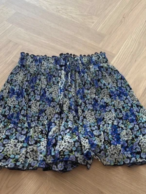 Blommig skort med smock och volang - Supersöt skort med blått, svart och vitt blommönster, smockad midja och volangkant. Skortsen har inbyggda svarta shorts och är gjord i ett lätt och luftigt material som känns bekvämt. Perfekt för dig som gillar att sticka ut med mönster!
