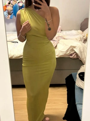 Lång limegrön oneshoulder-klänning - Snygg långklänning i limegrönt med oneshoulder-design och öppen rygg. Klänningen har en knytning bak och sitter tight över kroppen. Perfekt för dig som vill sticka ut med en unik och modern look. Materialet känns mjukt och följsamt.
