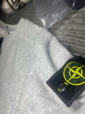 Grå, svart och vit tröja från Stone Island - Snygg sweatshirt från Stone Island med klassisk rund halsringning och ribbade muddar. Tröjan har en enkel en stilren design, perfekt för dig som gillar att sticka ut och exklusiva märken. Materialet känns mjukt och bekvämt mot huden.
