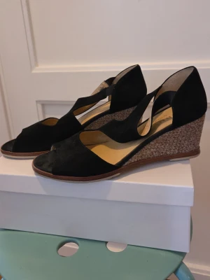 Svarta kilklack-sandaler med flätad sula - Snygga svarta sandaler med öppen tå och kilklack. Ovandelen är i mocka och klacken har en flätad detalj som ger en cool kontrast. Perfekt för dig som vill ha både stil och komfort. Klacken är medelhög och sulan har en brun kant. Medföljer även tillhörande skopåse.