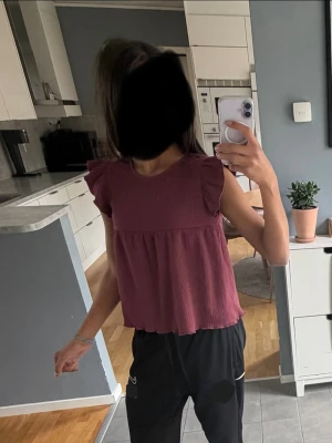 Vinröd ärmlös topp med volang - Säljer en vinröd ärmlös topp med volangdetaljer på axlarna. Toppen har en lös passform och är tillverkad i ett mjukt, strukturerat tyg som känns luftigt och skönt. Perfekt för dig som gillar en avslappnad men söt stil.