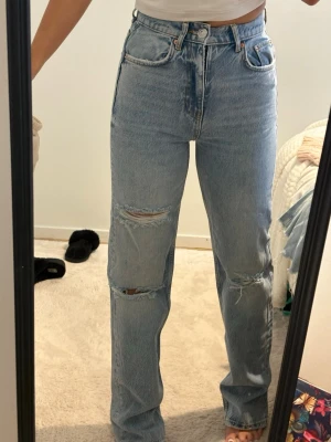 Ljusblå raka jeans med slitningar - Säljer ett par ljusblå jeans med hög midja och raka ben. Jeansen har slitna detaljer och hål på båda knäna för en trendig, avslappnad look. Klassisk femficksmodell och stängs med knapp och dragkedja. Perfekta till vardagsoutfits.