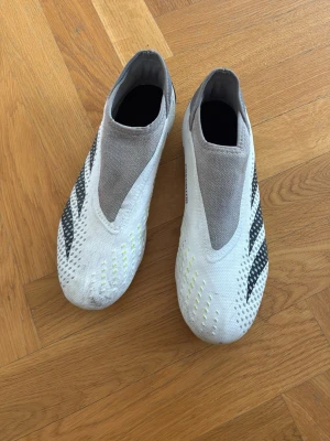 Adidas Predator vita fotbollsskor - Säljer ett par Adidas Predator fotbollsskor i vitt med svarta detaljer och neon-gula inslag. Skorna har en strumpliknande design utan snörning och är tillverkade i syntetmaterial för optimal passform och grepp på planen. Perfekta för dig som vill ha kontroll och precision.