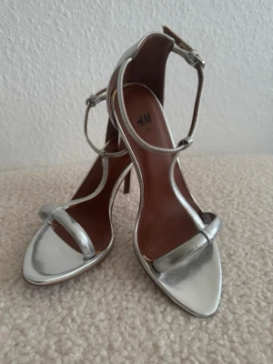 Nya. Silvriga högklackade sandaler H&M - Säljer ett par stilrena silvriga sandaler från H&M med smala remmar och öppen tå. Skorna har en elegant hög klack och justerbar ankelrem. Perfekta för dig som vill ha en modern och glansig look. Materialet är syntet med metallicfinish. Aldrig använd, men eftersom skorna stod 2 år i garderoben, så finns små skavanker, som inte är synliga, när man har skorna på, se sista 2 bilder, därav priset på 100kr.