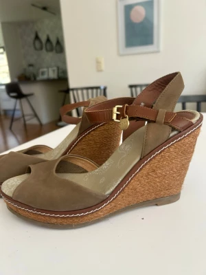 Bruna kilklackade sandaler från mexx - Snygga bruna sandaler från mexx med kilklack i flätad jute och öppen tå. Remmar i läder med guldfärgat spänne och dekorativ liten medaljong. Mjukt mönstrat innersula och vit söm längs kanten. Perfekt för sommarens outfits.