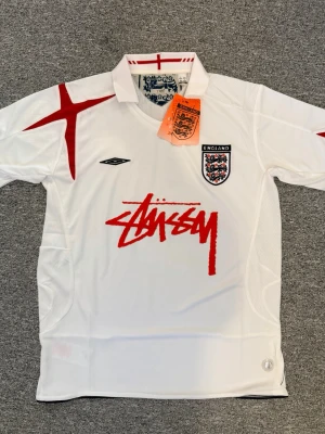 England x Stüssy vit fotbollströja - Custom vit fotbollströja från Stüssy i samarbete med Englands och Umbro. Den är helt ny, 68cm på längden och 51 på brädden. Hör av dig med frågor och behov av fler bilder!🤗