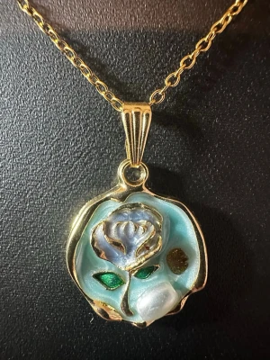 Guldigt halsband med blomma - Unikt halsband med tunn guldfärgad kedja och ett runt hänge i guldton. Hänget har ett vackert blommotiv med inslag av ljusblått och grönt emaljliknande material. Perfekt för dig som gillar detaljer och vill ha något som sticker ut.