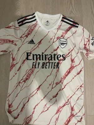 Arsenal bortatröja Adidas röd/vit - Säljer en Arsenal bortatröja från Adidas i vitt med röda marmorerade linjer. Tröjan har svarta detaljer, klubbmärke på bröstet och 'Emirates Fly Better' tryckt framtill. Tillverkad i lätt och ventilerande material, perfekt för match eller träning.