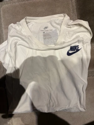 Vit Nike T-shirt med stor logga - Säljer en vit T-shirt från Nike med klassisk blå logga på bröstet och en stor Nike-tryck i blått över ryggen. T-shirten har korta ärmar och är gjord i mjuk bomull, perfekt för en sportig och avslappnad stil.