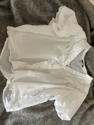 Vit blus med spetsdetaljer - Söt vit blus med korta puffärmar och fina spetsdetaljer längs knäppningen och halsringningen. Tröjan är perfekt till sommaren, jag säljer denna blus eftersom den inte passar mig😔