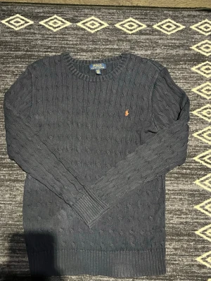 Mörkblå stickad tröja Polo Ralph Lauren - Snygg mörkblå kabelstickad tröja från Polo Ralph Lauren med klassisk orange logga broderad på bröstet. Tröjan har rund halsringning, långa ärmar och ribbade muddar. Perfekt för dig som gillar stilrena och tidlösa plagg.