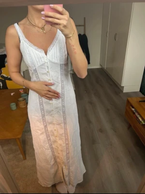 Vit långklänning med spetsdetaljer - Säljer en vit långklänning med smala axelband, v-ringning och dekorativa spetsdetaljer längs knappraden framtill. Klänningen har en feminin och bohemisk känsla, knappar hela vägen ner och är tillverkad i ett lätt tyg som passar perfekt till varma dagar.