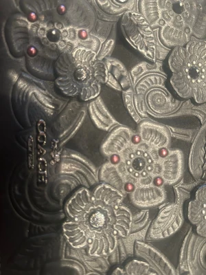 Svart plånbok med blommönster Coach - Säljer en svart plånbok från Coach i äkta läder med detaljerat blommönster i relief och små silver- och roséfärgade nitar. Plånboken har tryckknapp, dragkedjefack och flera fack för kort. Märket Coach är präglat både på utsidan och insidan.