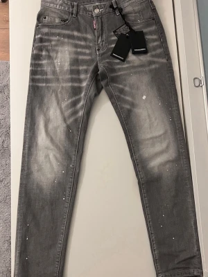 Grå Dsquared2 jeans med stänk - Säljer ett par grå jeans från Dsquared2 med vita färgstänk och slitningar. Jeansen har rak passform, fem fickor och patch med 'Caten Heated' bak. Tillverkade i denim med cool streetkänsla och detaljer som sticker ut.