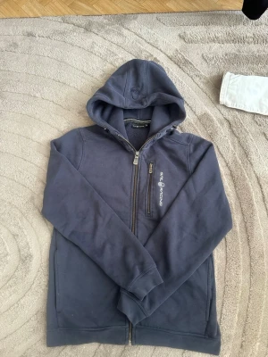 Marinblå hoodie från Sail Racing - Snygg marinblå hoodie från Sail Racing med dragkedja framtill och en extra bröstficka med dragkedja. Hoodien har broderad logga på bröstet och en skön huva. Perfekt för chill dagar eller när du vill ha en sportig look.