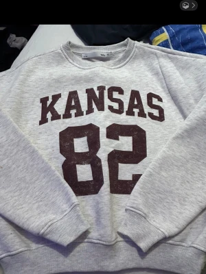 Grå sweatshirt med KANSAS 82-print - Säljer en grå sweatshirt  med stort vinrött tryck där det står KANSAS och siffran 82 på framsidan. Tröjan har rund halsringning, ribbade muddar vid ärmslut och nederkant. Perfekt för en avslappnad och sportig stil.
