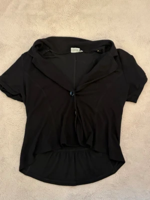 Svart kortärmad figursydd topp - Svart topp med korta ärmar och en figursydd modell. Toppen har en bred krage och är något croppad baktill. Perfekt att styla över ett linne eller klänning för en cool lager-på-lager-look.