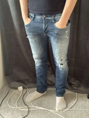 Blå replay anbass hyperflex jeans - Säljer ett par blå skinny jeans med slitningar och hål på låret och knät. Jeansen har en tvättad look med ljusare partier och klassisk femficksdesign. Perfekta för en avslappnad och trendig stil.