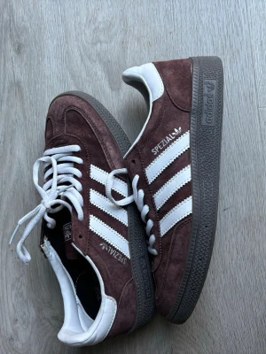 Adidas Spezial bruna sneakers - Säljer ett par Adidas Spezial sneakers i mörkbrunt mocka med vita detaljer och klassiska tre ränder på sidan. Skorna har vita skosnören och en grå sula. Märket och modellen syns tydligt på sidan. Perfekta för dig som gillar retrostil och streetwear. (Viktigt - dem är inte äkta)