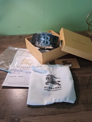 Svart Burberry bälte med silverdetalj - Snyggt svart bälte från Burberry med en stor, silverfärgad B-logga som spänne. Bältet kommer i originalförpackning med dustbag och certifikat. Perfekt accessoar för att lyfta din outfit med en exklusiv touch.