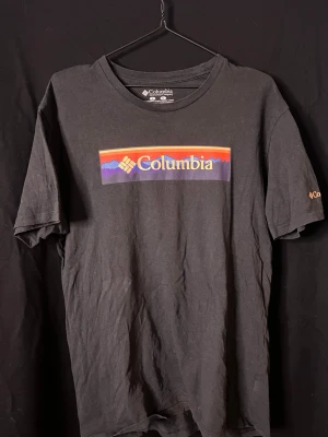 Svart Columbia t-shirt med tryck - Säljer en svart t-shirt från Columbia med färgglatt tryck av logga och berg på bröstet. Klassisk rund hals och korta ärmar. Tillverkad i 100% bomull, perfekt för en avslappnad och sportig stil.