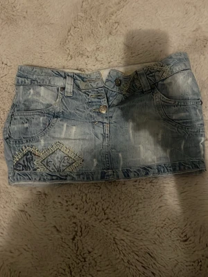 Ljusblå jeanskjol med stjärntecken broderi - Säljer en kort ljusblå jeanskjol med slitna detaljer och broderier av vädurs tecknet framtill. Kjolen har fram och bakfickor, och stängs med knappar, en knapp är tyvärr trasig i mitten som man kan se på bilden. Jag köpte den på second hand, och säljer den pga att den inte passar mig. ;(