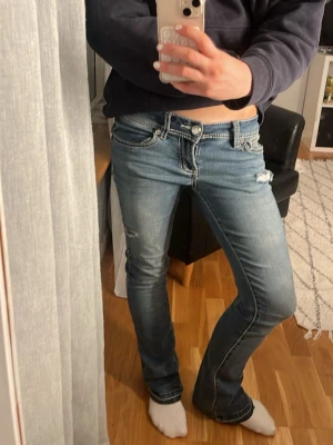 Blå bootcut jeans med broderad ficka - Säljer ett par blå bootcut jeans med slitningar och snygga kontrastsömmar. Bakfickan har ett detaljerat broderi med silvertråd och nitar. Jeansen har låg midja och klassisk femficksmodell. Perfekta för dig som gillar en lite mer edgy stil. Litet hål vid fig ändan som inte märks av