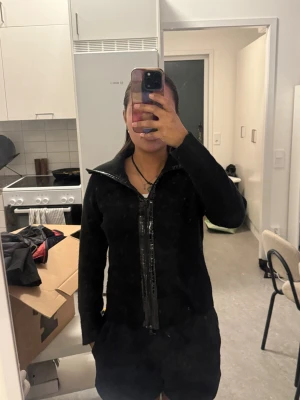 Svart kofta med dragkedja och detaljer - Säljer en svart kofta med dragkedja framtill och glänsande detaljer längs dragkedjan. Hoodien har lång ärm och en hög krage som kan vikas ner. Perfekt för dig som gillar en stilren men ändå unik look. Storlek mellan xs och S