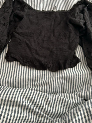 Svart croppad topp med spetsärmar - Trendig svart croppad topp med långa spetsärmar och smockad kropp. Toppen har volangkant nedtill och en snygg offshoulder-look. Perfekt för dig som vill ha en stilren men ändå detaljrik topp till din outfit.