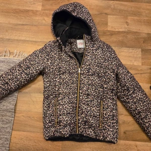Leopardmönstrad pufferjacka med huva - Säljer en trendig leopardmönstrad pufferjacka med huva och dragkedja framtill. Jackan har två fickor med guldfärgade dragkedjor och svart innerfoder. Perfekt för dig som vill sticka ut i vinterkylan.