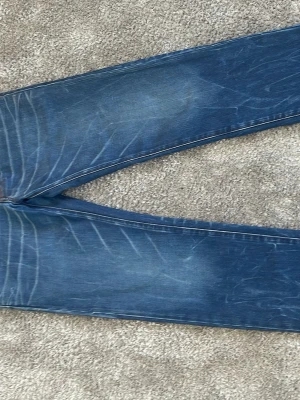 Blå slim fit jeans med slitningar - Snygga blå slim fit jeans med tydliga slitningar och whiskers-effekter på både fram- och baksida. Klassisk femficksmodell med kontrastsömmar och läderpatch bak i midjan. Perfekta för en avslappnad och trendig look.  Str 31/32