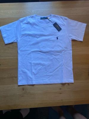 Vit t-shirt från Polo Ralph Lauren - Klassisk vit t-shirt från Polo Ralph Lauren med rund halsringning och korta ärmar. T-shirten har den ikoniska mörkblå broderade loggan på bröstet och är tillverkad i mjuk bomull för en skön känsla. Perfekt basplagg till garderoben.