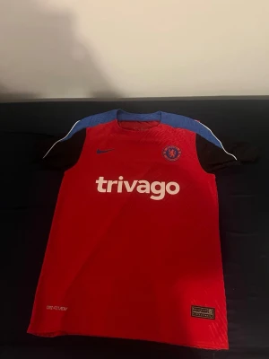 Chelsea röd tränings t-shirt Nike - Chelsea FC tränings t-shirt i rött med svarta ärmar och blå axelparti. Tröjan har vit trivago-logga på bröstet, Chelsea-märke och Nike-logga. Tillverkad i lätt och snabbtorkande material med Dri-FIT-teknologi. Perfekt för fotbollsträning.