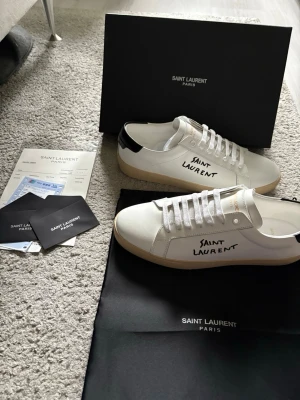 Saint Laurent vita sneakers med svarta detaljer - Säljer ett par vita Saint Laurent sneakers i läder med svart detalj på hälen och handskriven logga på sidan. Skorna har vita snören, gummisula och klassisk låg profil. Perfekta för dig som gillar stilrena och exklusiva sneakers.