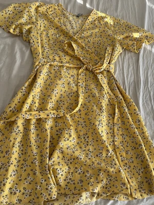 Gul blommig omlottklänning - Söt gul omlottklänning med små blommor i vitt och svart. Klänningen har korta ärmar, knytband i midjan och volangdetaljer längs nederkanten. Perfekt för soliga dagar och ger en fräsch sommarkänsla.