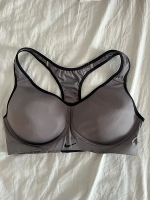 Grå sport-bh från Nike - Sport-bh i grått med svarta detaljer från Nike. Bh:n har vadderade kupor och brottarrygg för extra stöd och komfort. Tillverkad i ett stretchigt funktionsmaterial som andas. Perfekt för träning och aktiva dagar.