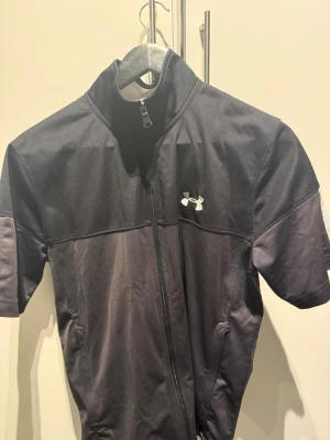 Svart träningsjacka Under Armour - Snygg svart träningsjacka från Under Armour med korta ärmar och hel dragkedja framtill. Jackan har en hög krage och det klassiska Under Armour-loggan på bröstet. Perfekt för träning eller löpning. Tillverkad i ett lätt och funktionellt material.