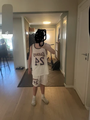 Lakers basketset med nummer 24 - Säljer ett vitt basketset med Lakers-tryck och nummer 24 i lila och guld. Setet består av ett linne och matchande shorts, båda i meshmaterial som andas. Perfekt för basketträning eller streetstyle. Klassisk sportig look med kontrastfärgade kanter.