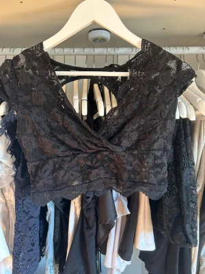 Svart spetsig croppad topp - Snygg svart croppad topp i spets med omlott framtill och öppen rygg. Toppen har fina blommiga spetsdetaljer och knappar bak. Perfekt för dig som vill ha en trendig och unik look.