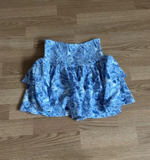 Blå blommig volangkjol Gina Tricot  - Säljer en söt blå kortkjol med blommigt mönster och volanger. Kjolen har en bred, smockad midja som ger en bekväm passform och extra snygg siluett. Perfekt för dig som gillar romantiska och somriga vibes.